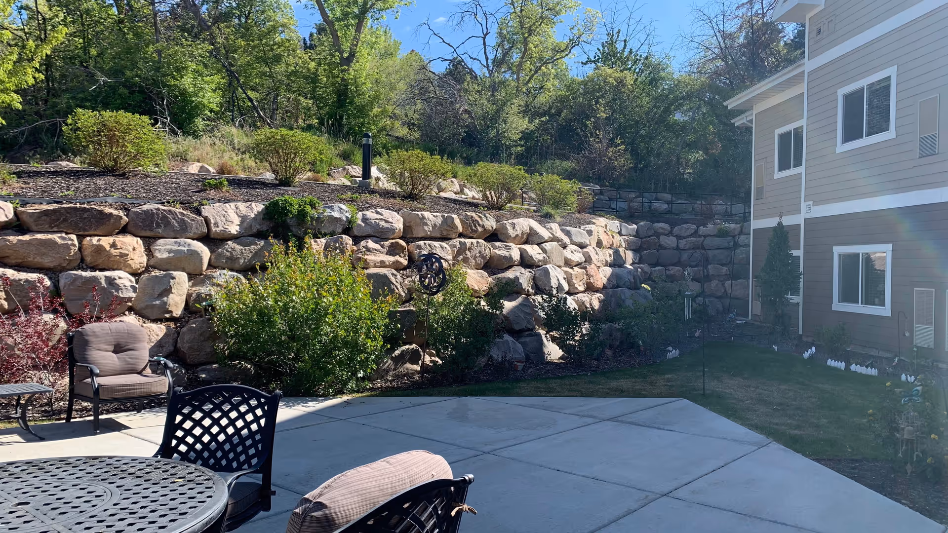 Outdoor patio area with metal chairs and a round table, surrounded by greenery and a stone retaining wall. A beige building with white trim is visible on the right side under a clear blue sky.