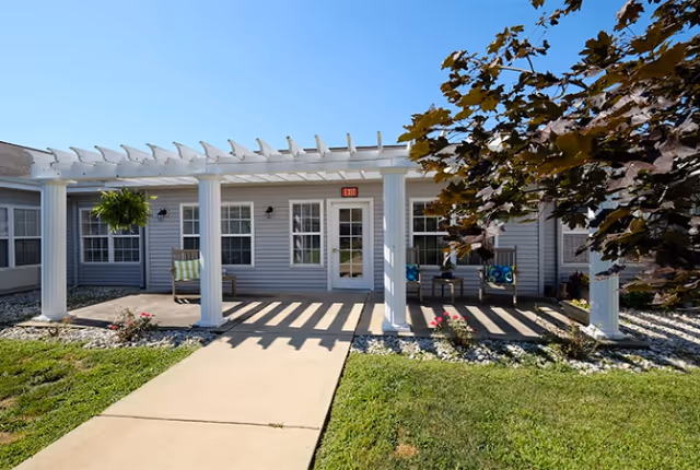 Outdoor patio area at Digby Place featuring a white pergola with columns, a concrete walkway leading to a door with an exit sign, hanging plants, and outdoor seating with cushions. There is a tree with dark leaves partially shading the area and a well-maintained lawn surrounding the patio.