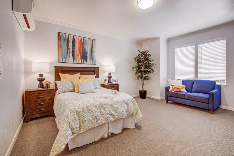 A bright and spacious bedroom with a neatly made bed featuring multiple pillows and a patterned comforter. There are two wooden nightstands with lamps and small decorative items on either side of the bed. A colorful abstract painting hangs above the headboard. To the right, a blue sofa with decorative pillows sits in front of three windows with white blinds. A tall potted plant is placed in the corner of the room. The walls are white, and the floor is carpeted in a neutral tone.
