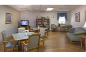 A cozy senior living lounge with tables, chairs, a sofa, bookshelf, and a television.