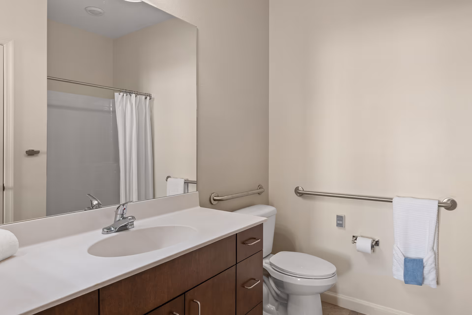 A clean bathroom with a white countertop and sink, a large mirror above the sink, a toilet with grab bars on the walls, a towel rack with a white towel and a blue washcloth, and a shower with a white curtain.
