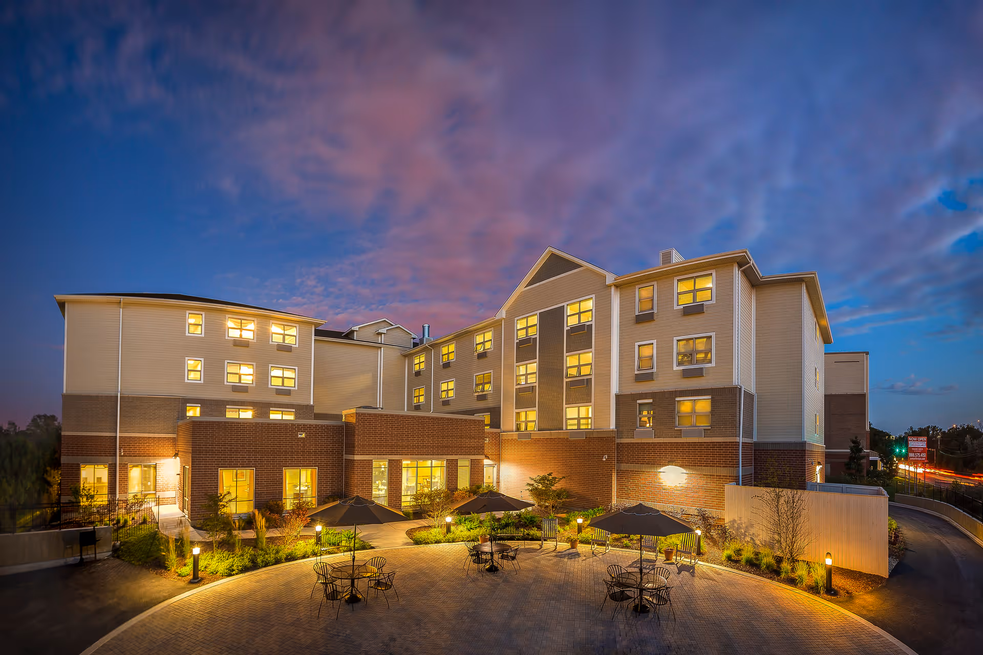 Evening view of a multi-story senior living facility building with warm interior lights glowing through the windows. In front of the building is a paved courtyard with several round tables and chairs under large umbrellas, surrounded by landscaped greenery and illuminated by small outdoor lamps. The sky is a mix of blue and purple hues with scattered clouds.