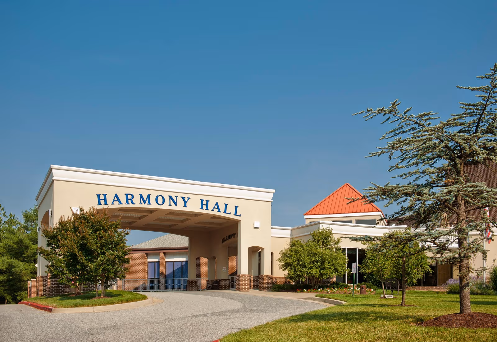 Exterior view of Harmony Hall building with a covered entrance, surrounded by trees and greenery under a clear blue sky.