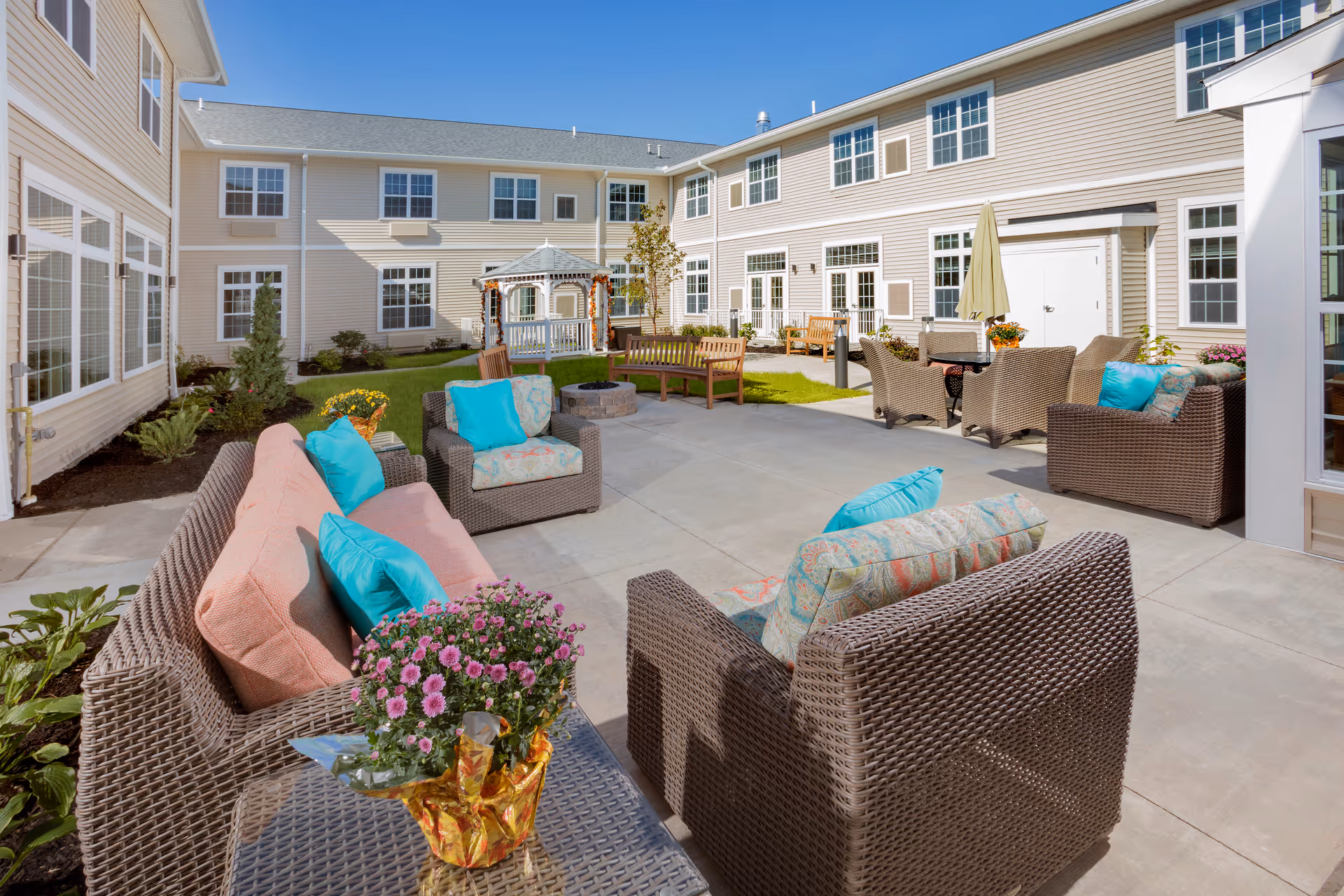 Sunny outdoor courtyard with wicker seating, colorful cushions, potted flowers, and a gazebo surrounded by a two-story building.