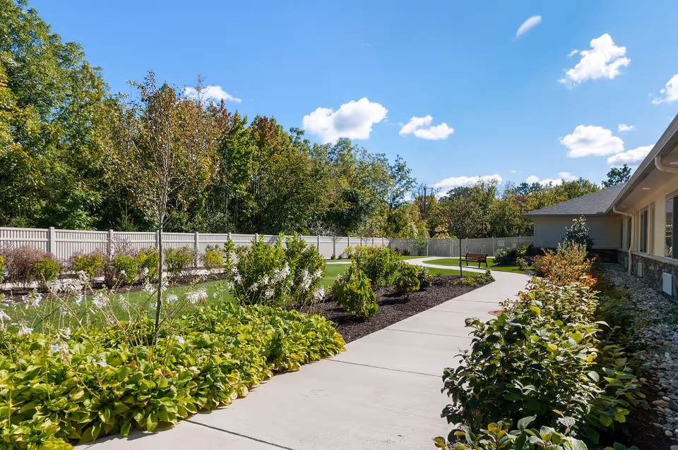 A sunny outdoor garden area at Viva Memory Care at Dresher featuring a curved concrete walkway bordered by green shrubs and plants. There is a wooden bench along the path and a tall fence enclosing the garden. Trees with green and some autumn-colored leaves are visible in the background under a blue sky with scattered white clouds.