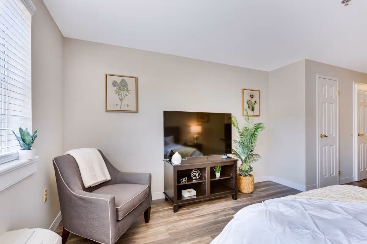 A cozy bedroom corner featuring a gray armchair with a white throw draped over it, a dark wooden TV stand with a flat-screen television, decorative items, and a potted plant in a woven basket. The room has light-colored walls, two framed botanical prints, and wood flooring. A bed with white bedding is partially visible in the foreground.