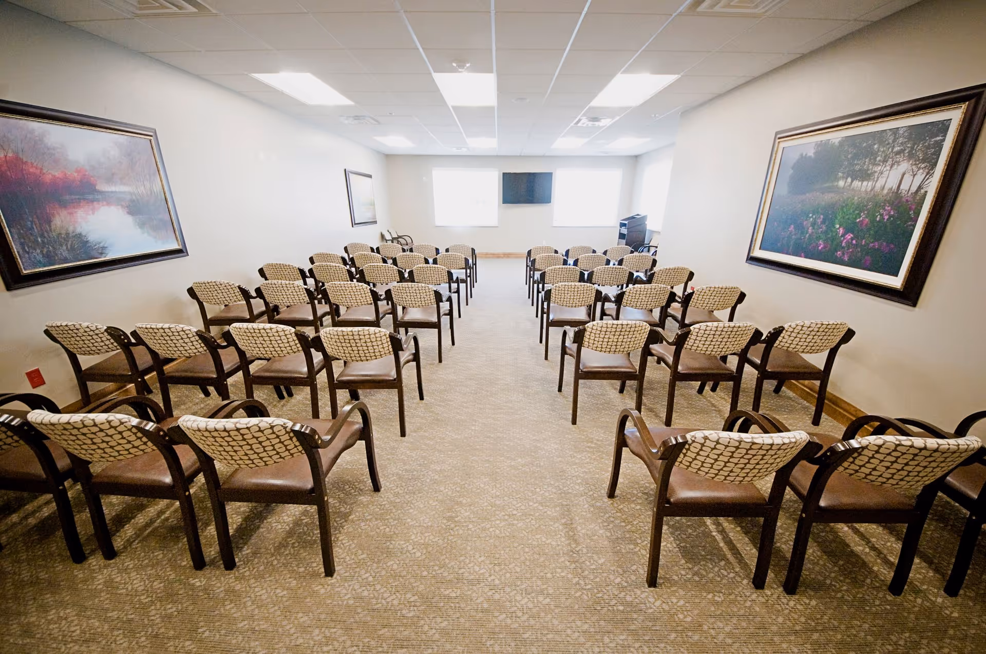 Rows of chairs arranged theater-style facing a wall-mounted screen and podium in a well-lit meeting room with framed artwork.