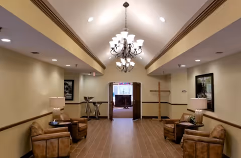 A hallway with beige walls and brown tiled floor, featuring four brown leather armchairs arranged in pairs on either side. Each pair has a side table with a lamp. A chandelier hangs from the ceiling, and double doors at the end of the hallway lead to a room with a cross and stained glass window.