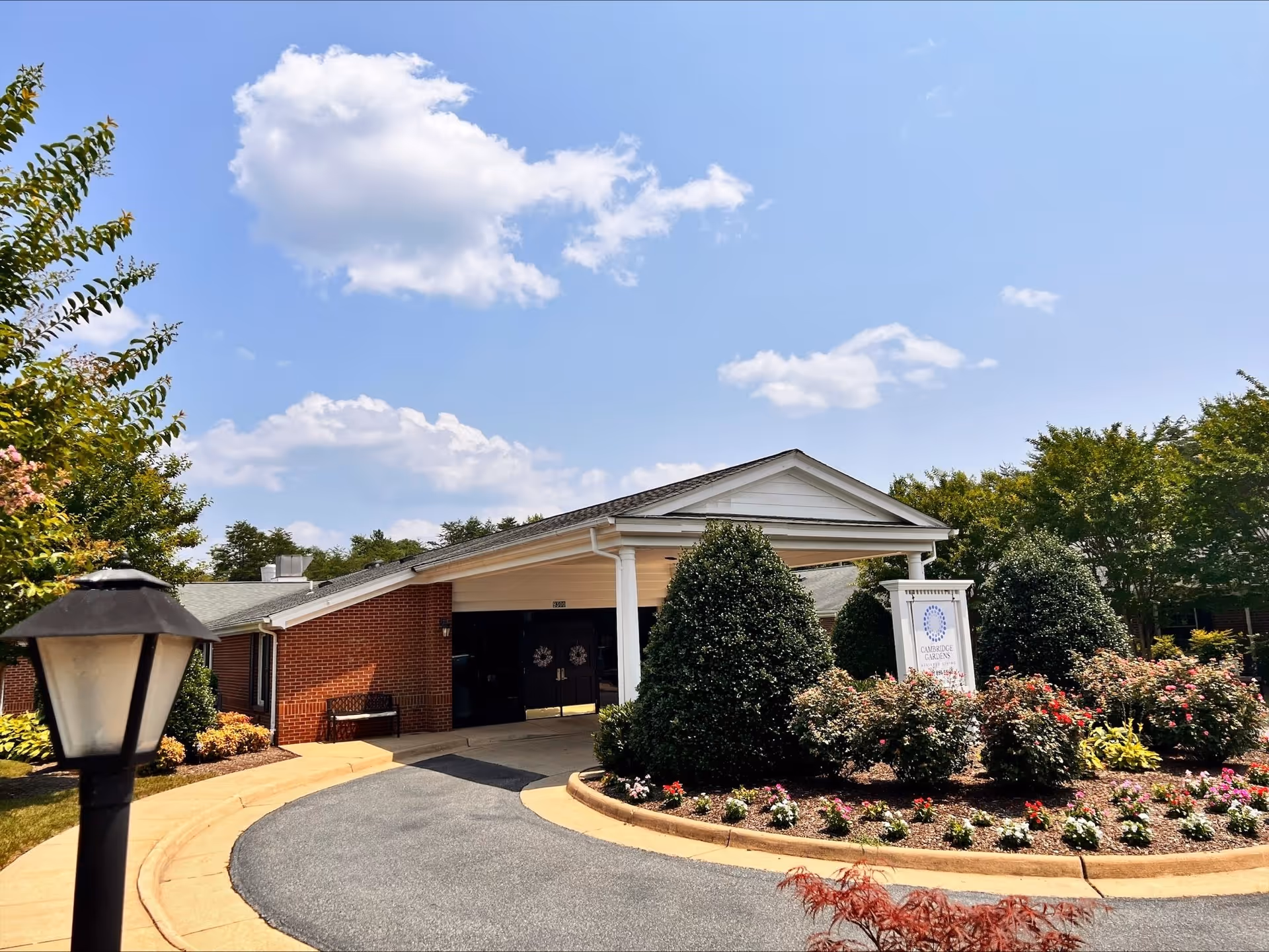 Exterior view of a single-story brick building with a covered entrance and a circular driveway. The building is surrounded by well-maintained landscaping including bushes, flowers, and trees under a blue sky with scattered clouds.