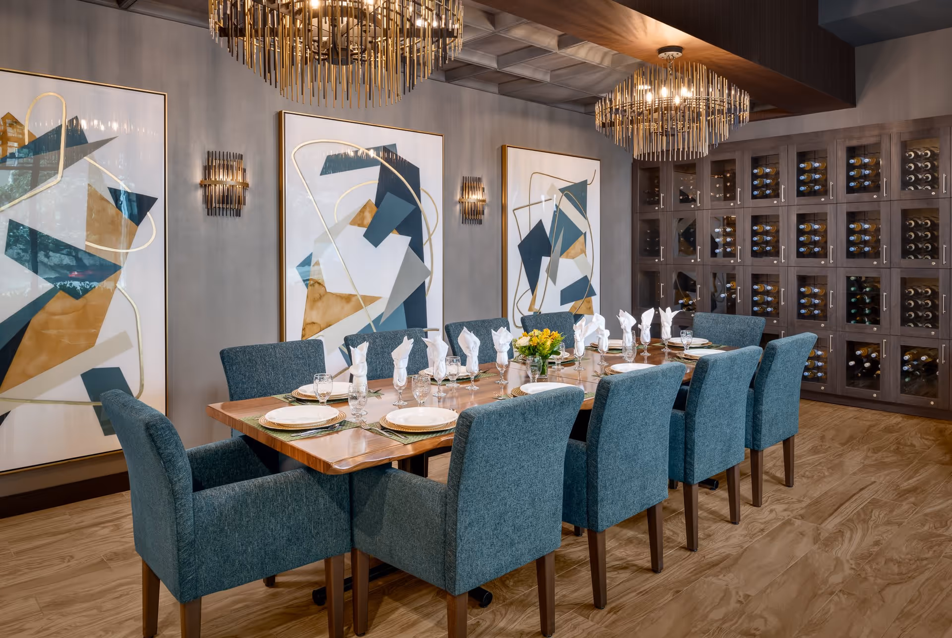 A modern dining room with a long wooden table set for ten people with white plates, glasses, and folded napkins. The chairs are upholstered in teal fabric. The room features three large abstract paintings on the wall, two elegant chandeliers, and a large wine rack filled with bottles.