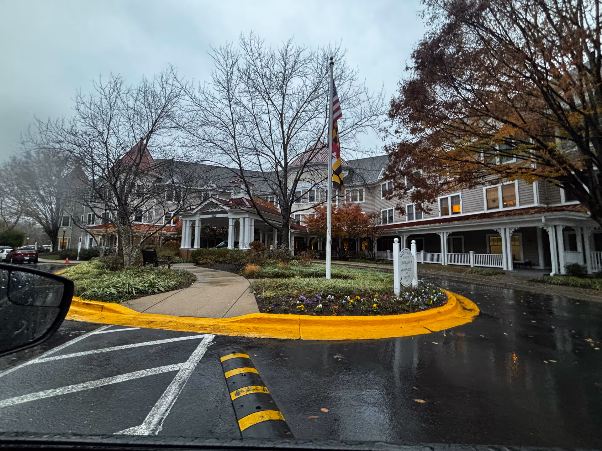 Exterior view of a senior living facility building with a covered entrance, a flagpole with the American and Maryland flags, landscaped garden beds, and trees with autumn foliage on a rainy day.