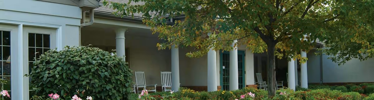 Exterior view of a senior living facility with white columns, a covered porch with rocking chairs, surrounded by green bushes, trees, and flowering plants.