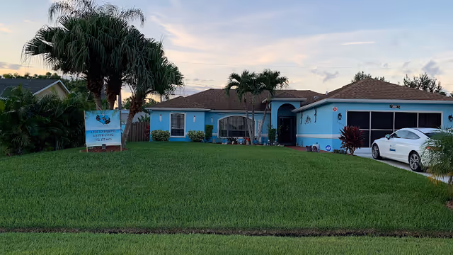 A single-story blue building with a brown roof, surrounded by palm trees and green grass under a partly cloudy sky at sunset. A white car is parked in the driveway on the right side. A sign on the lawn reads 'A Kee To Paradise Assisted Living' with a phone number.