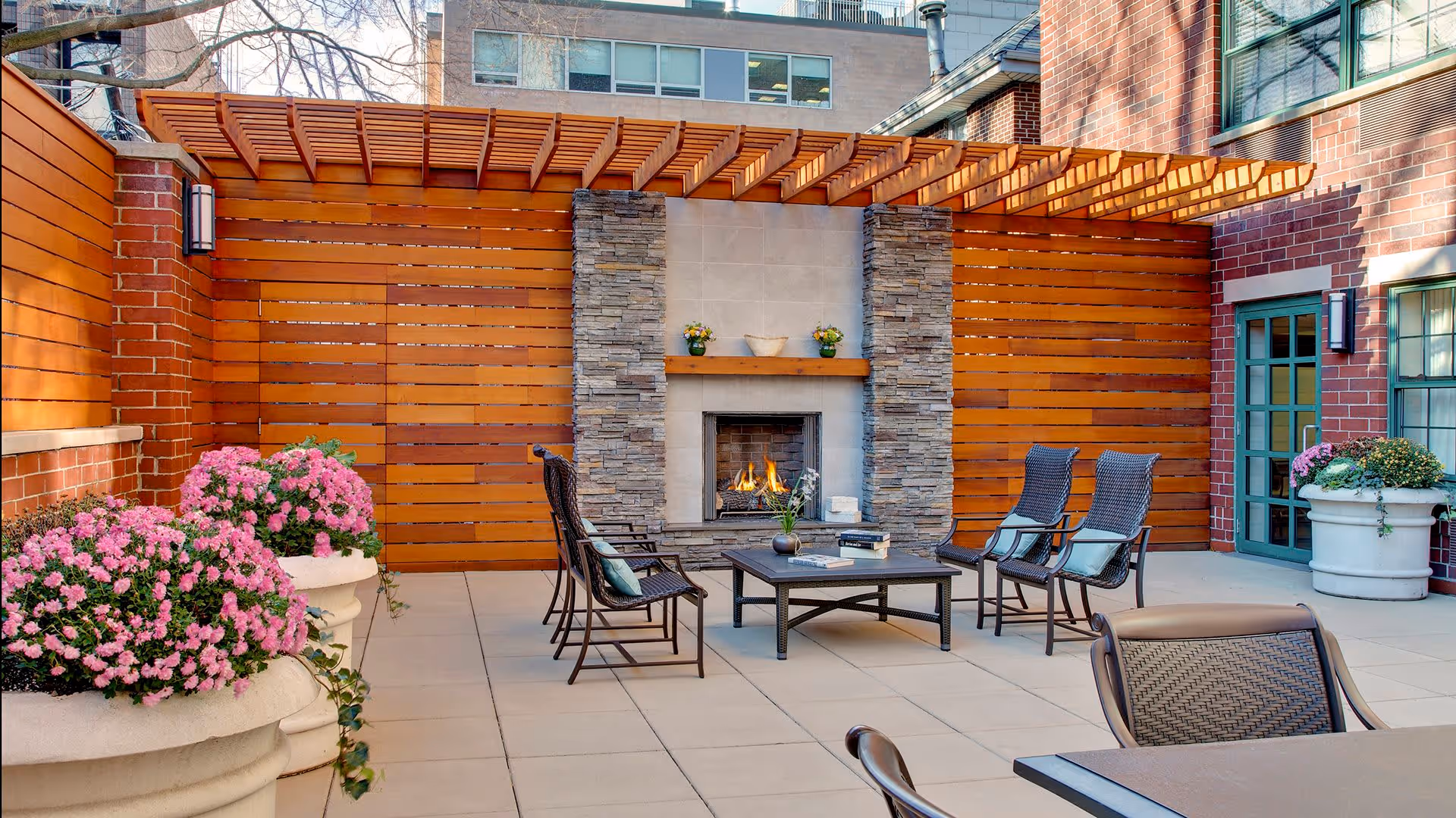 Outdoor patio area with a stone fireplace surrounded by a wooden pergola and wooden privacy walls. There are several black metal chairs with cushions arranged around a low coffee table. Large planters with pink flowers and greenery are placed around the patio. The area is adjacent to a brick building with windows and a door.