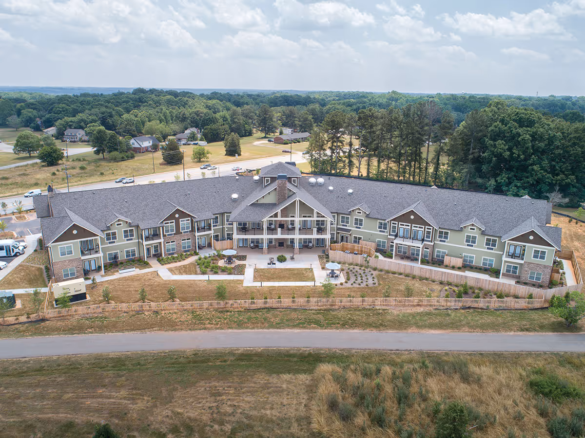 Aerial view of a large senior living facility building with a gray roof and light green exterior walls. The building has multiple windows and balconies, with a fenced outdoor patio area featuring tables and umbrellas. Surrounding the facility are trees, grassy fields, and a road in the foreground.