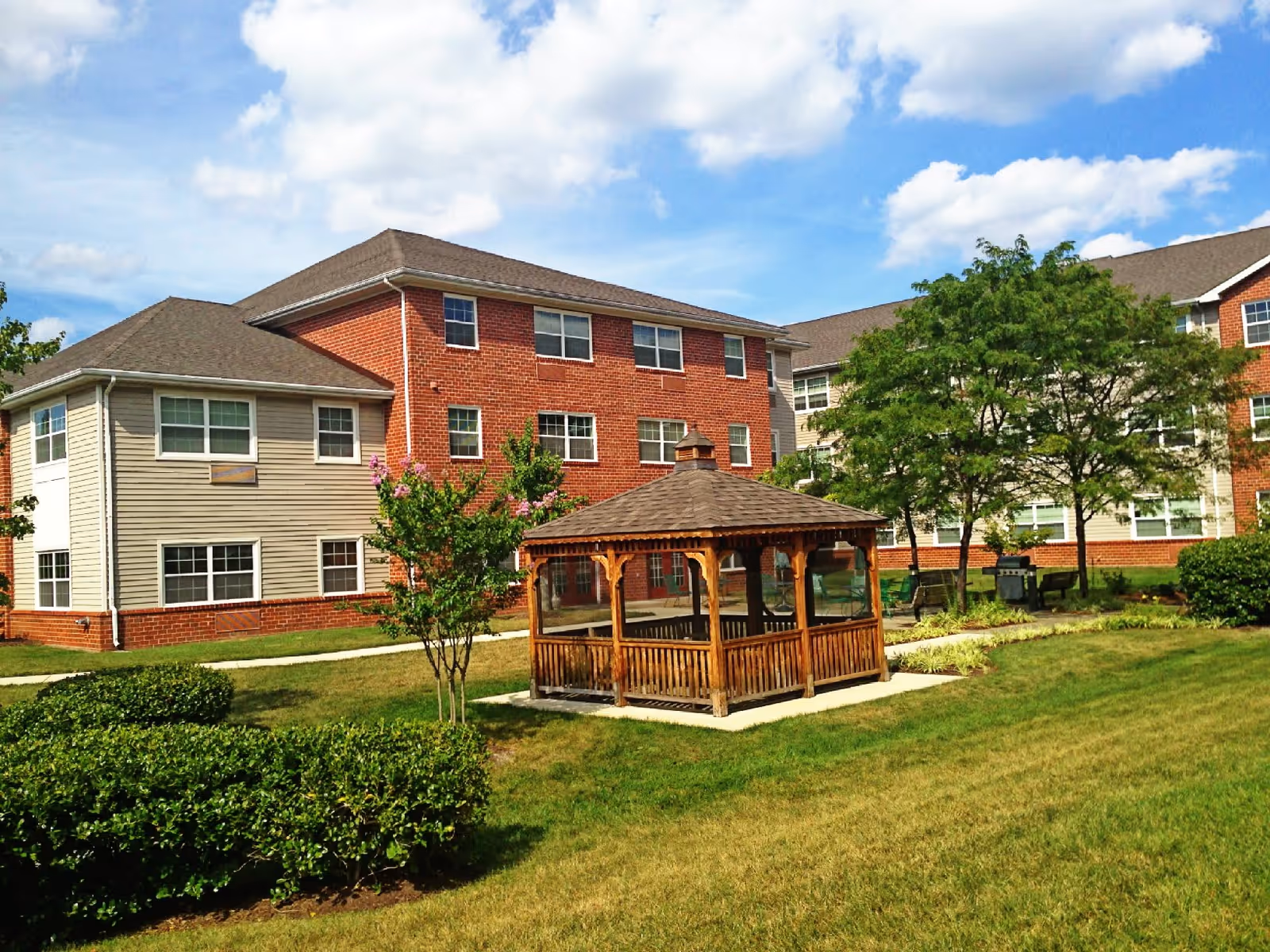 Outdoor view of a senior living facility with a wooden gazebo in the center, surrounded by green grass, bushes, and trees. The background shows a multi-story building with red brick and beige siding under a partly cloudy blue sky.