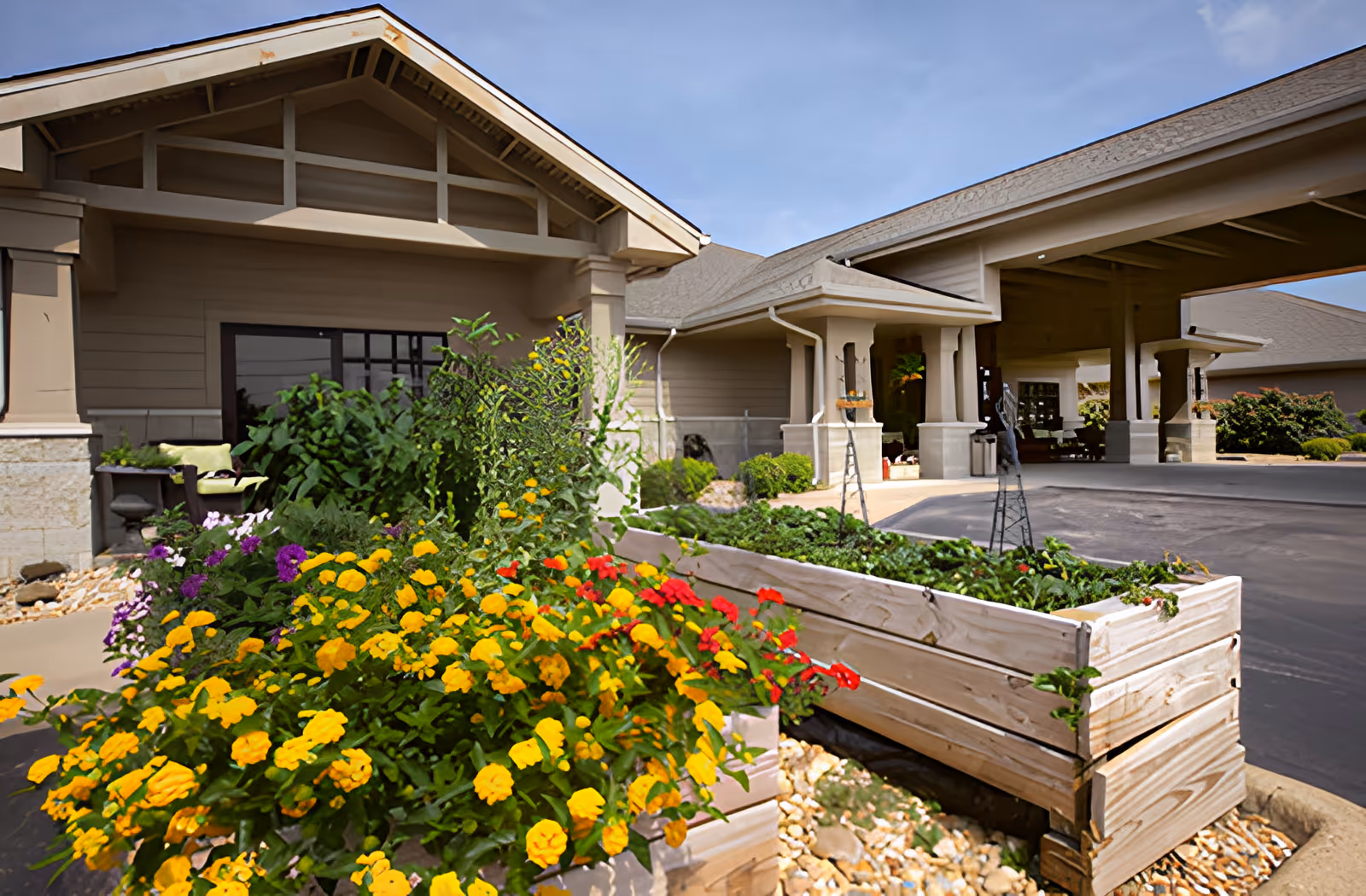 Exterior view of Ahc West Tennessee Transitional Care facility showing a covered entrance with pillars, a driveway, and raised garden beds filled with colorful flowers and greenery in the foreground.