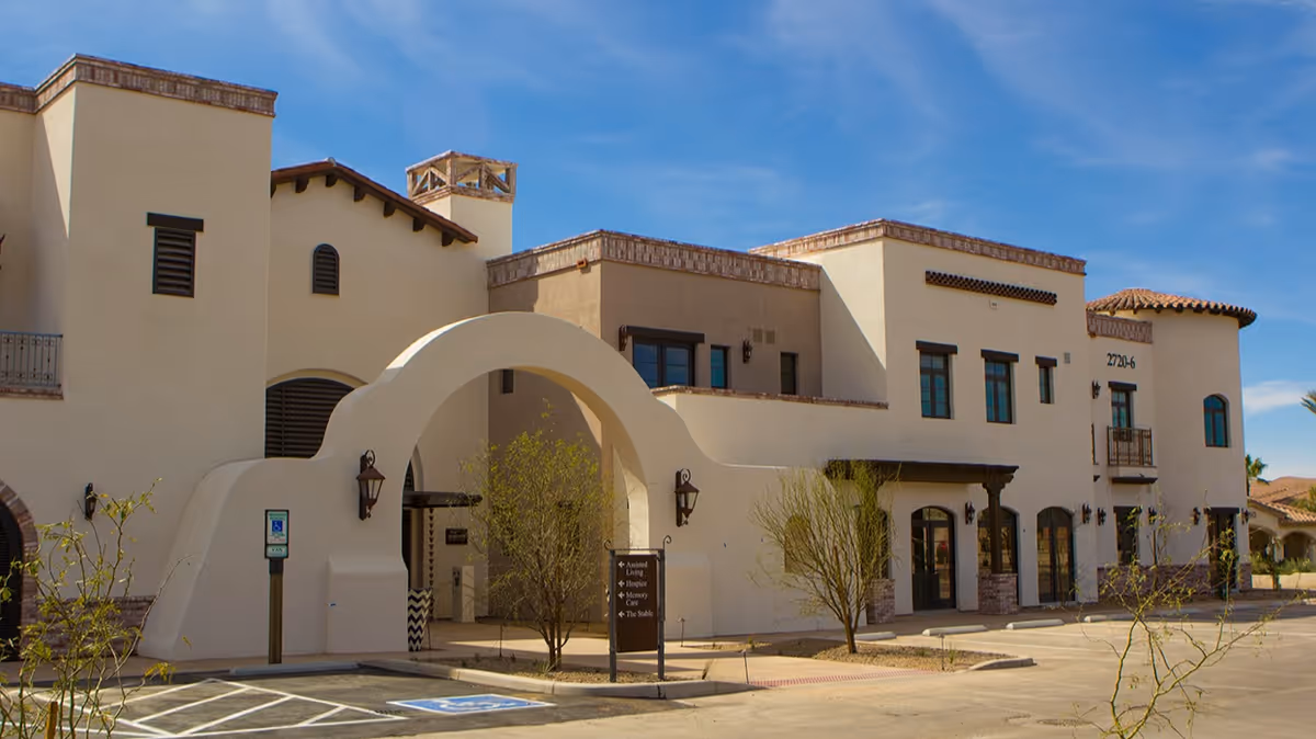 Exterior view of a large beige building with Spanish-style architecture under a clear blue sky. The building features an arched entrance, multiple windows, and a small balcony. There are a few small trees and a parking area with a handicap parking space in front.