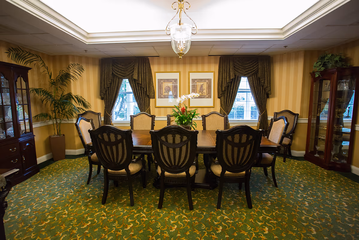 A formal dining room with a long wooden table surrounded by upholstered chairs, patterned carpet, draped windows, and glass-front china cabinets.