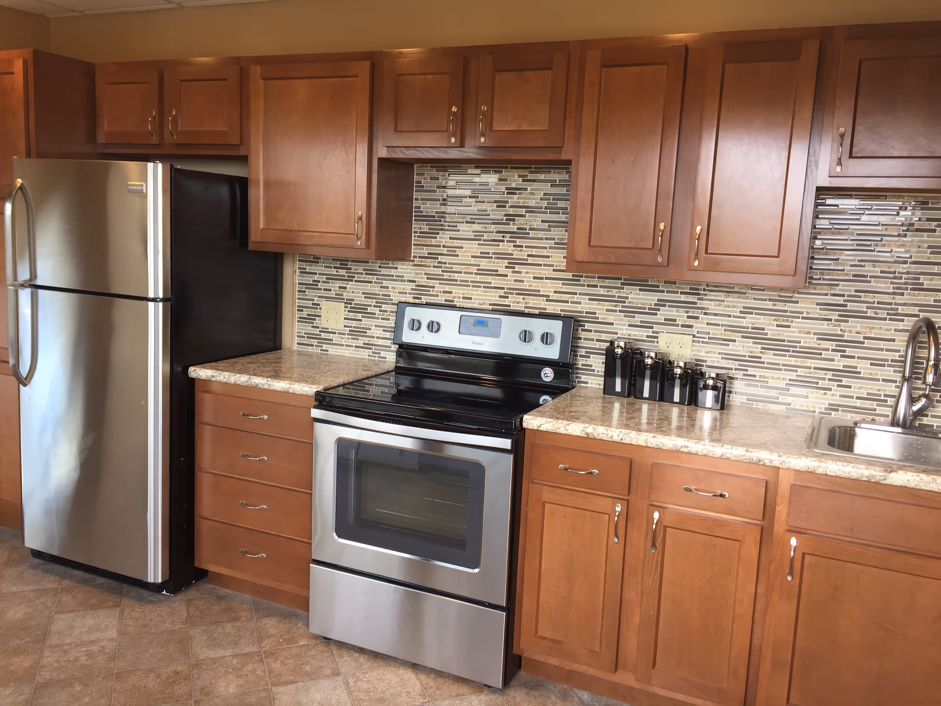 A modern kitchen featuring wooden cabinets, a stainless steel refrigerator, an electric stove with oven, a tiled backsplash in neutral tones, a countertop with several black canisters, and a stainless steel sink with a faucet.