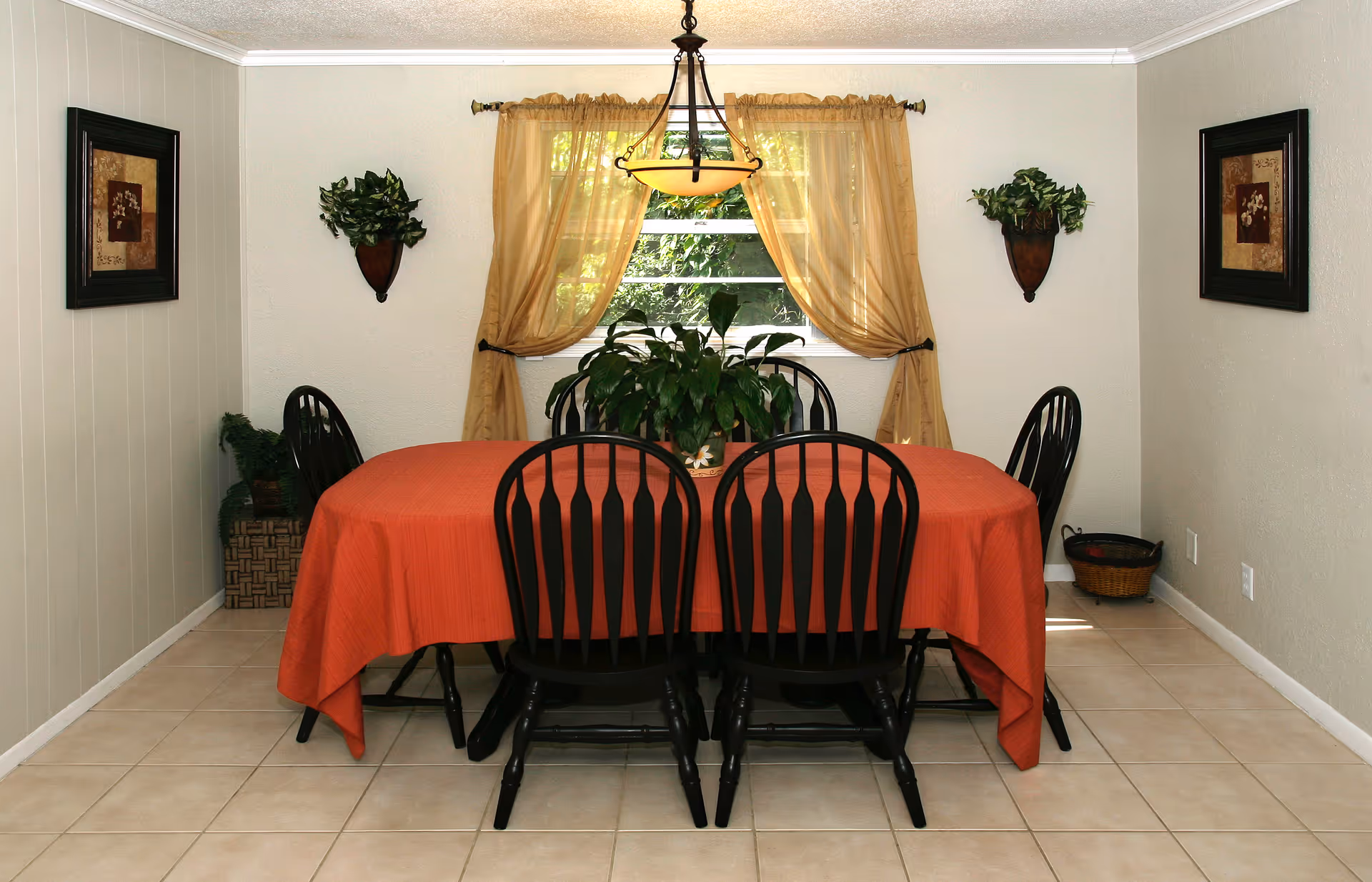 A dining room with a rectangular table covered with an orange tablecloth and six black wooden chairs. A green potted plant is centered on the table. The room has beige tiled flooring, light-colored walls, and a window with sheer beige curtains tied back. Two framed floral artworks hang on the walls, and two wall-mounted planters with green foliage are on either side of the window. A hanging light fixture is above the table.