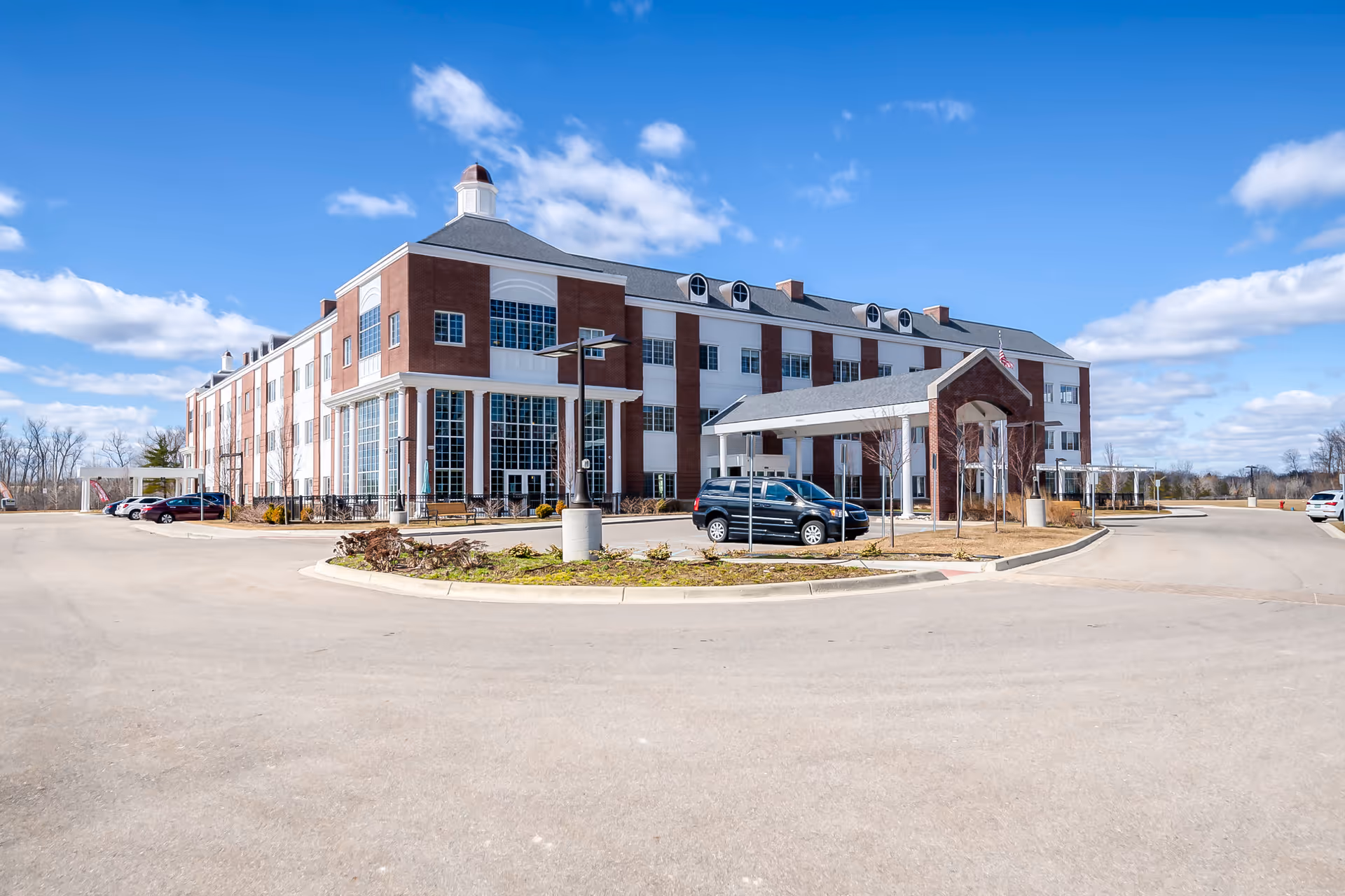 Front view of a three-story brick senior living building with a covered entrance and parked cars under a bright blue sky.