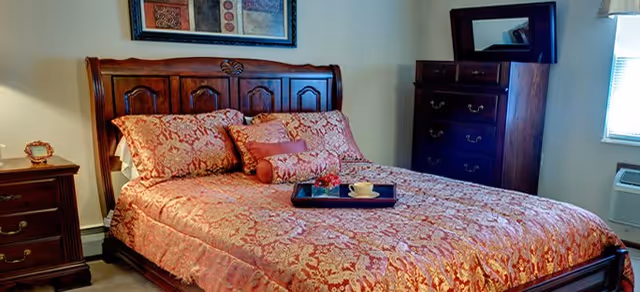 A bedroom with a large wooden bed featuring an ornate headboard. The bed is covered with a red and gold patterned bedspread and matching pillows. A dark wooden nightstand with a small lamp and decorative item is beside the bed. A dark wooden dresser with a mirror is positioned against the wall near a window with blinds. The room is softly lit and has a cozy, traditional style.