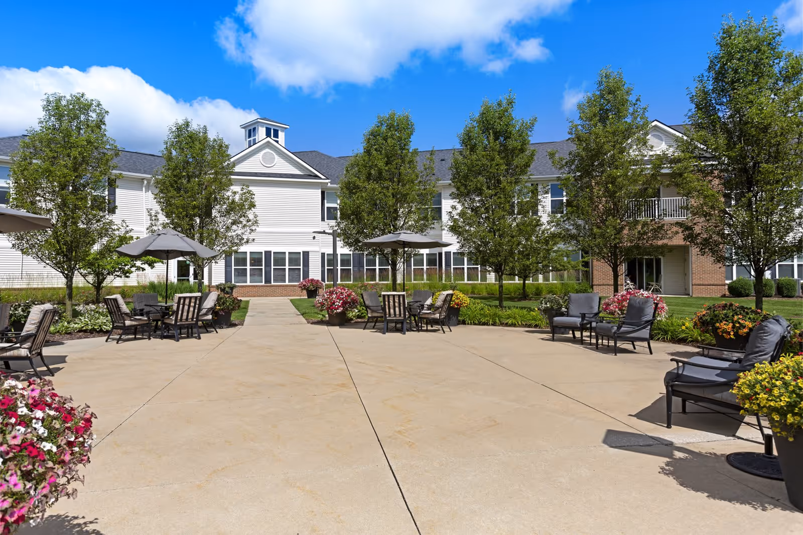 Outdoor patio area at Olivia's Assisted Living with multiple seating arrangements including chairs and tables with umbrellas, surrounded by trees and flower pots, and a two-story building in the background under a blue sky with some clouds.