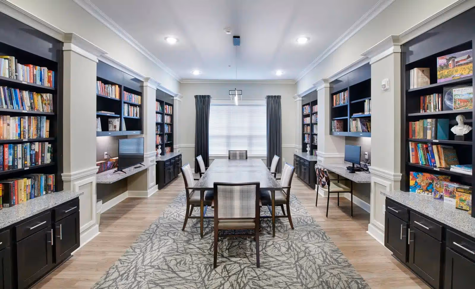 A well-lit room with a long rectangular table surrounded by eight chairs, flanked by built-in bookshelves filled with books and decorative items. The room has light-colored walls, a patterned carpet, and two computer workstations integrated into the bookshelves on either side. A large window with closed blinds and dark curtains is at the far end of the room.