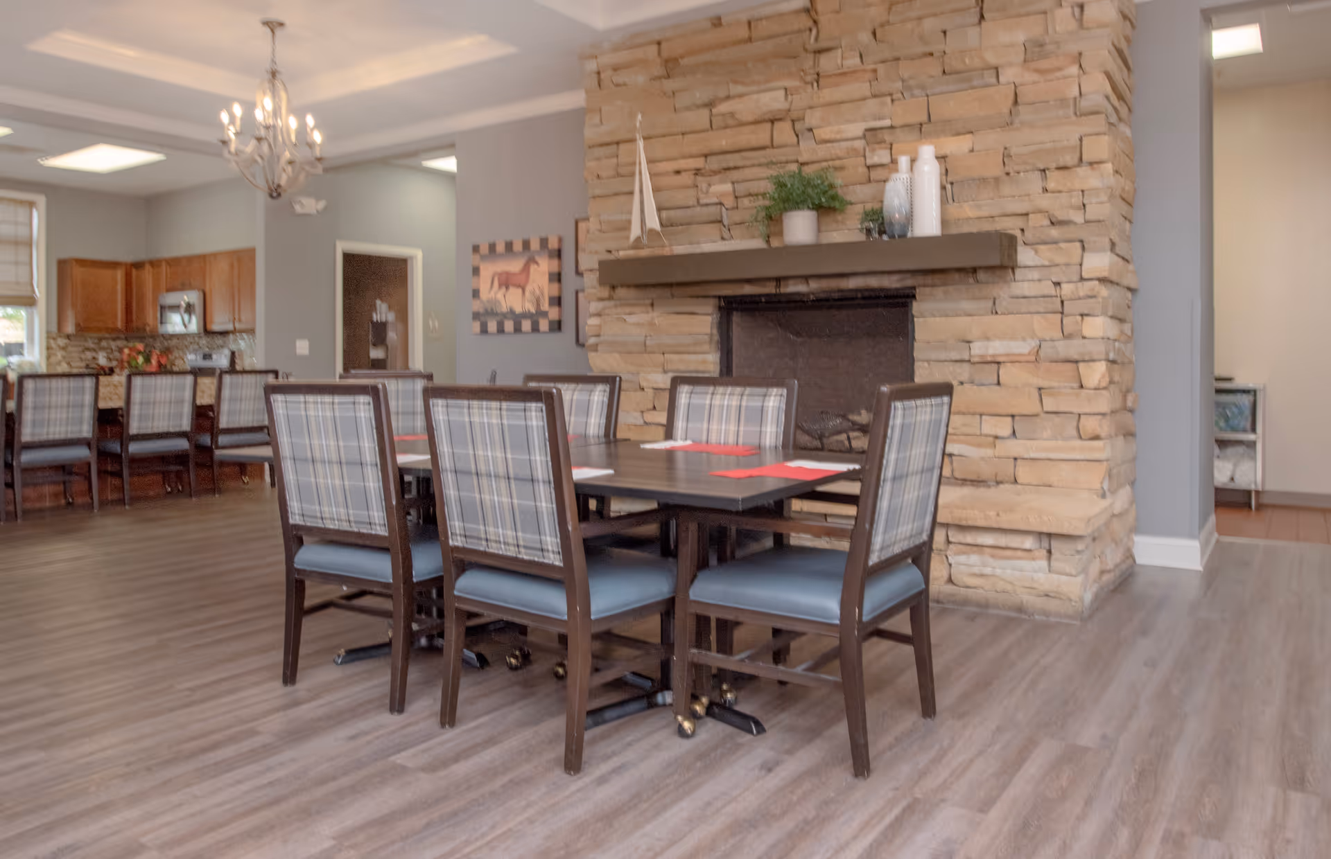 A dining area in a senior living facility featuring a dark wooden table surrounded by eight chairs with plaid backs and blue seats. Behind the table is a stone fireplace with decorative items on the mantel. To the left, there is a kitchen area with wooden cabinets and a countertop with several chairs. The room has wood flooring and soft gray walls, with a chandelier hanging from the ceiling.