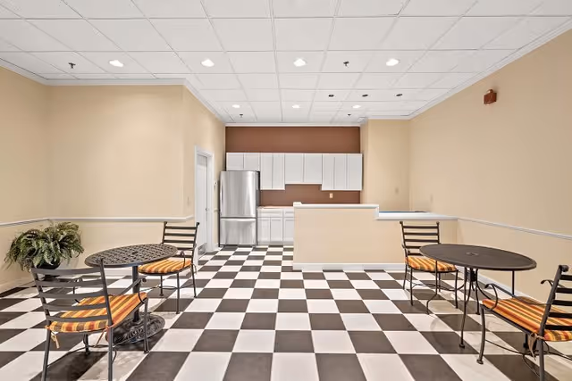 A small kitchen area with white cabinets and a stainless steel refrigerator at the back of a room with beige walls and a black and white checkered floor. In the foreground, there are two round black metal tables with striped orange and yellow cushioned chairs. A green potted plant is placed on the left side near the wall.