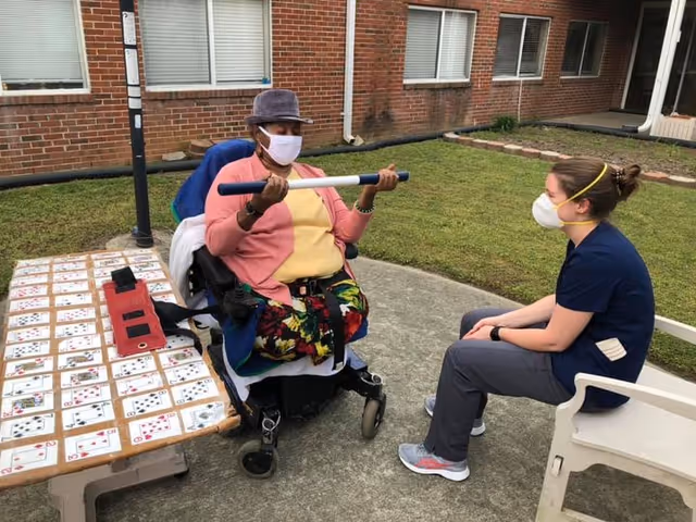 An elderly woman in a wheelchair wearing a hat and face mask is exercising with a bar while a caregiver, also wearing a face mask, sits on a chair in front of her outside a brick building. A table nearby is covered with playing cards and a red device.