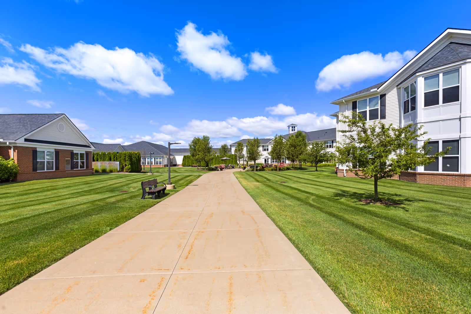 A wide concrete pathway runs through a well-maintained green lawn with small trees and benches on either side. Residential buildings with white siding and brick bases are visible on both sides under a bright blue sky with scattered white clouds.