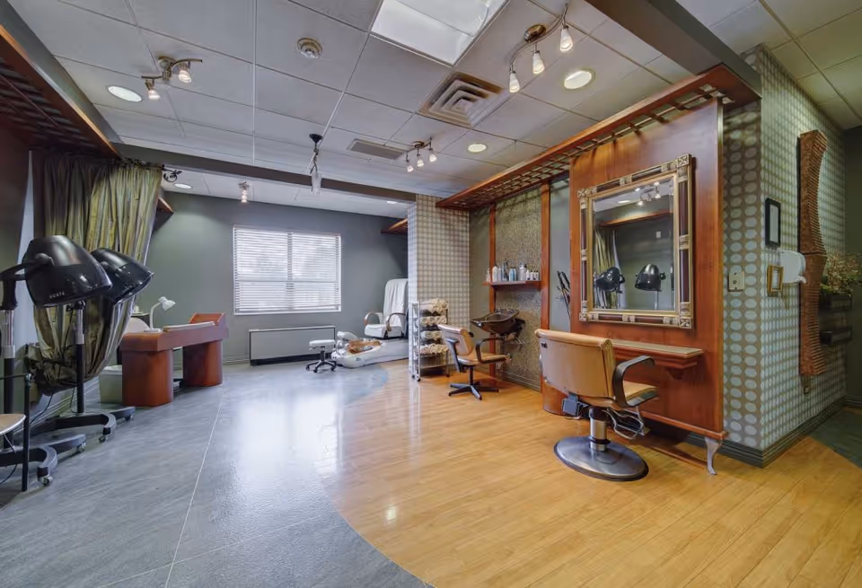 Interior view of a salon area in a senior living facility with hair drying stations, a large mirror, salon chairs, a pedicure chair, and shelves with towels and hair products. The room has a mix of wooden and tiled flooring and a window with blinds letting in natural light.