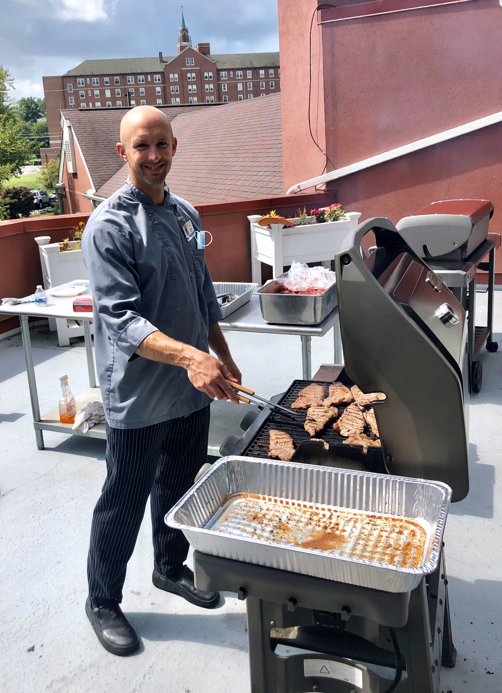 A man wearing a chef's coat and striped pants is grilling several pieces of meat on an outdoor grill on a rooftop patio. There are tables with trays and containers of food behind him, and a large brick building with a steeple is visible in the background.