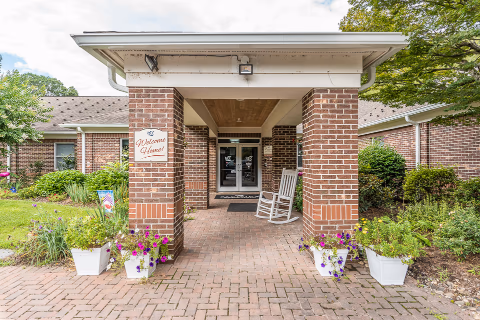 Entrance to a senior living facility with a covered brick walkway supported by brick columns. There are white rocking chairs and flower pots with colorful flowers on either side of the walkway. A sign on one of the columns reads 'Welcome Home!'. The building is surrounded by greenery and bushes.