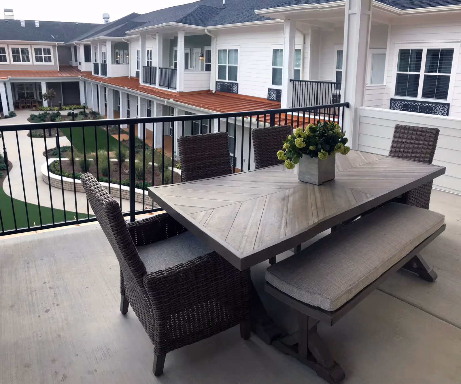 Outdoor balcony area with a rectangular wooden table, four wicker chairs, and a cushioned bench. A potted plant with green flowers is placed on the table. The balcony overlooks a courtyard with landscaped greenery and a two-story building with white siding and multiple windows.