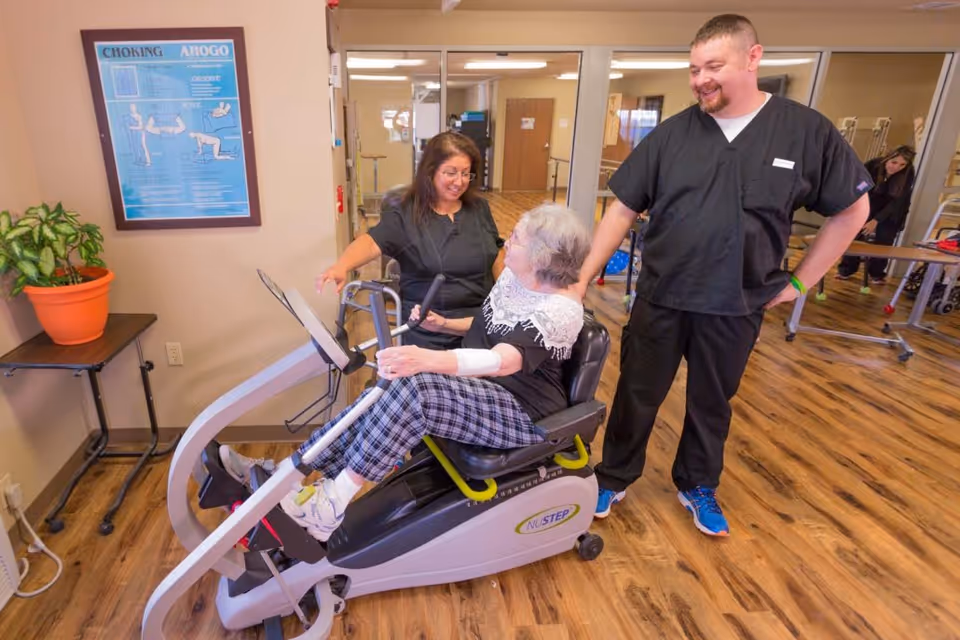 An elderly woman using a NuStep exercise machine in a rehabilitation facility, assisted by a female staff member and observed by a male staff member, both wearing black scrubs. The room has wooden flooring, a potted plant on a small table, and a framed choking first aid poster on the wall.