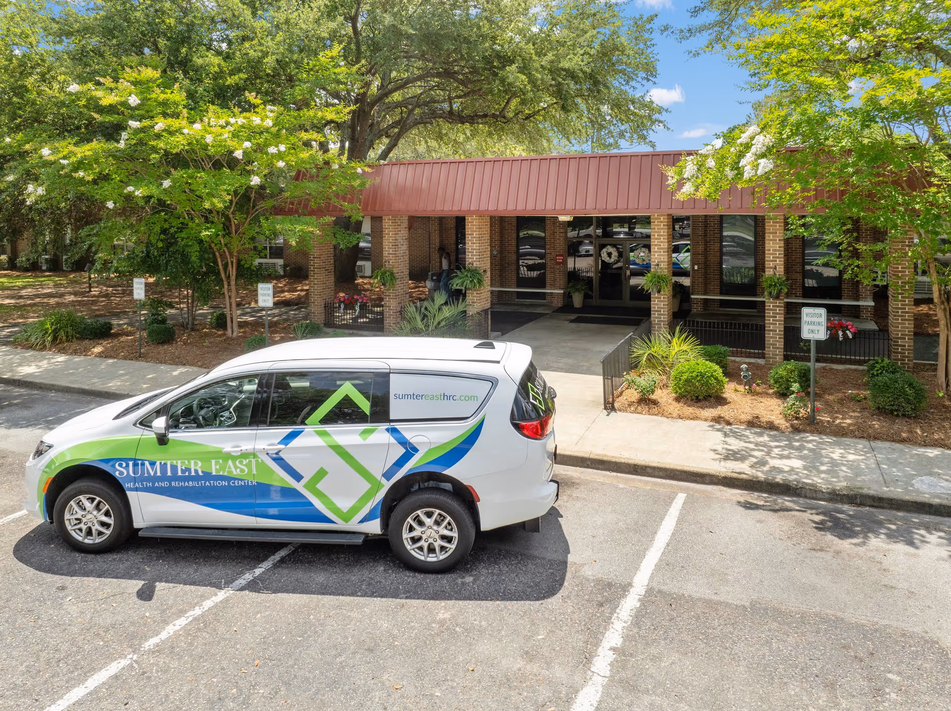 Exterior view of Sumter East Health and Rehabilitation Center showing the entrance with a covered walkway supported by brick columns. A white vehicle with the facility's logo and website is parked in front in a visitor parking spot. Trees and landscaping surround the entrance area under a clear blue sky.