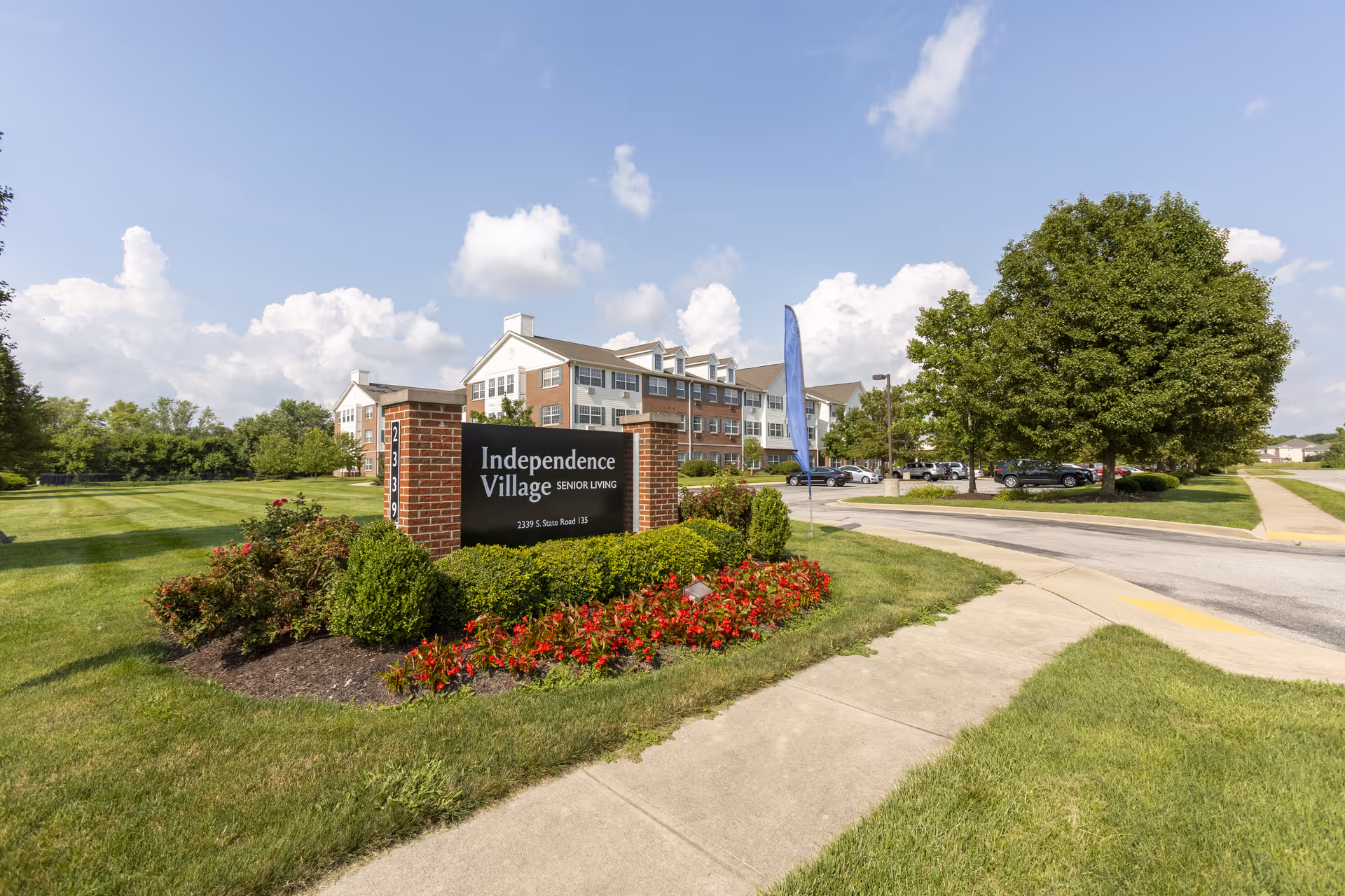 Exterior view of Independence Village Senior Living facility with a brick sign surrounded by flowers and bushes. The building is a multi-story structure with white and brick facade, set against a partly cloudy sky. There is a sidewalk and a parking area with cars visible.
