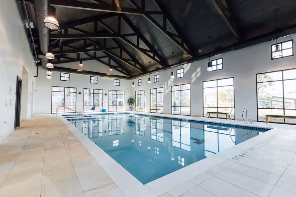 Indoor swimming pool with clear blue water, surrounded by large windows letting in natural light. The ceiling has exposed dark wooden beams and hanging lights. There are benches along the windows and a tiled floor around the pool.