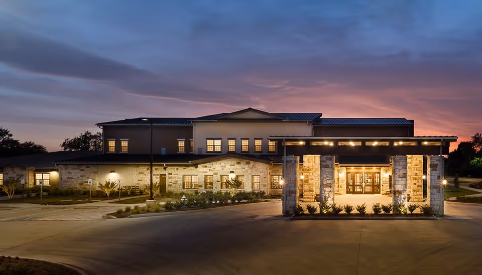 Exterior view of Spring Cypress Senior Living facility at dusk with illuminated entrance and a partly cloudy sky in the background.
