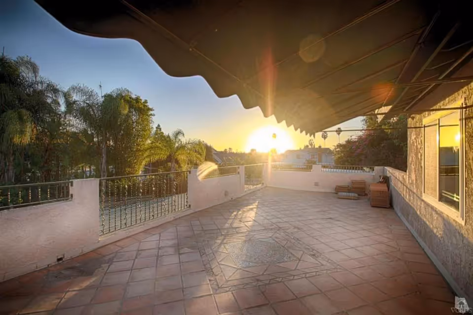 A spacious outdoor terrace with tiled flooring and a decorative tile pattern in the center. The terrace is bordered by a low wall with metal railings and overlooks palm trees and distant houses. The sun is setting, casting a warm glow over the scene. An awning extends over part of the terrace, and there are some stacked chairs and cushions near the wall.