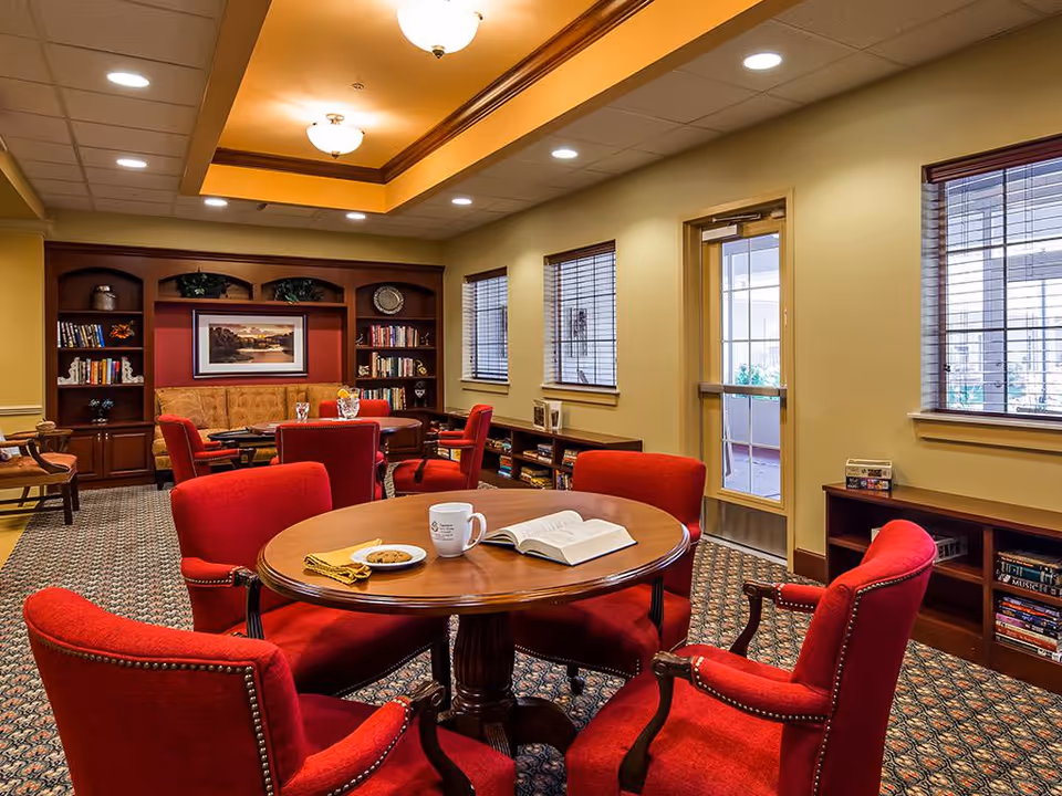 A cozy common area in a senior living facility featuring a round wooden table with four red upholstered chairs. On the table are a white mug, an open book, a plate with cookies, and a yellow napkin. The room has warm yellow walls, carpeted floor, and built-in wooden bookshelves filled with books and decorative items. There is a door and windows with blinds on one side, and a cushioned bench with more chairs and a framed landscape painting on the far wall.