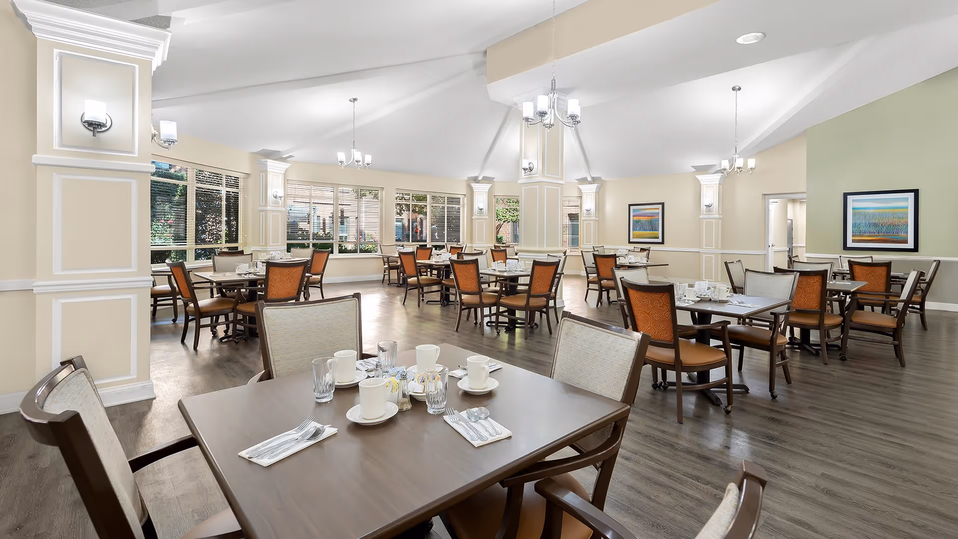 A spacious dining room with multiple tables and chairs arranged neatly. Each table is set with cups, glasses, napkins, and utensils. The room features large windows allowing natural light to brighten the space, light-colored walls with decorative molding, and framed artwork. The floor is wood, and there are several ceiling light fixtures and wall sconces providing additional lighting.