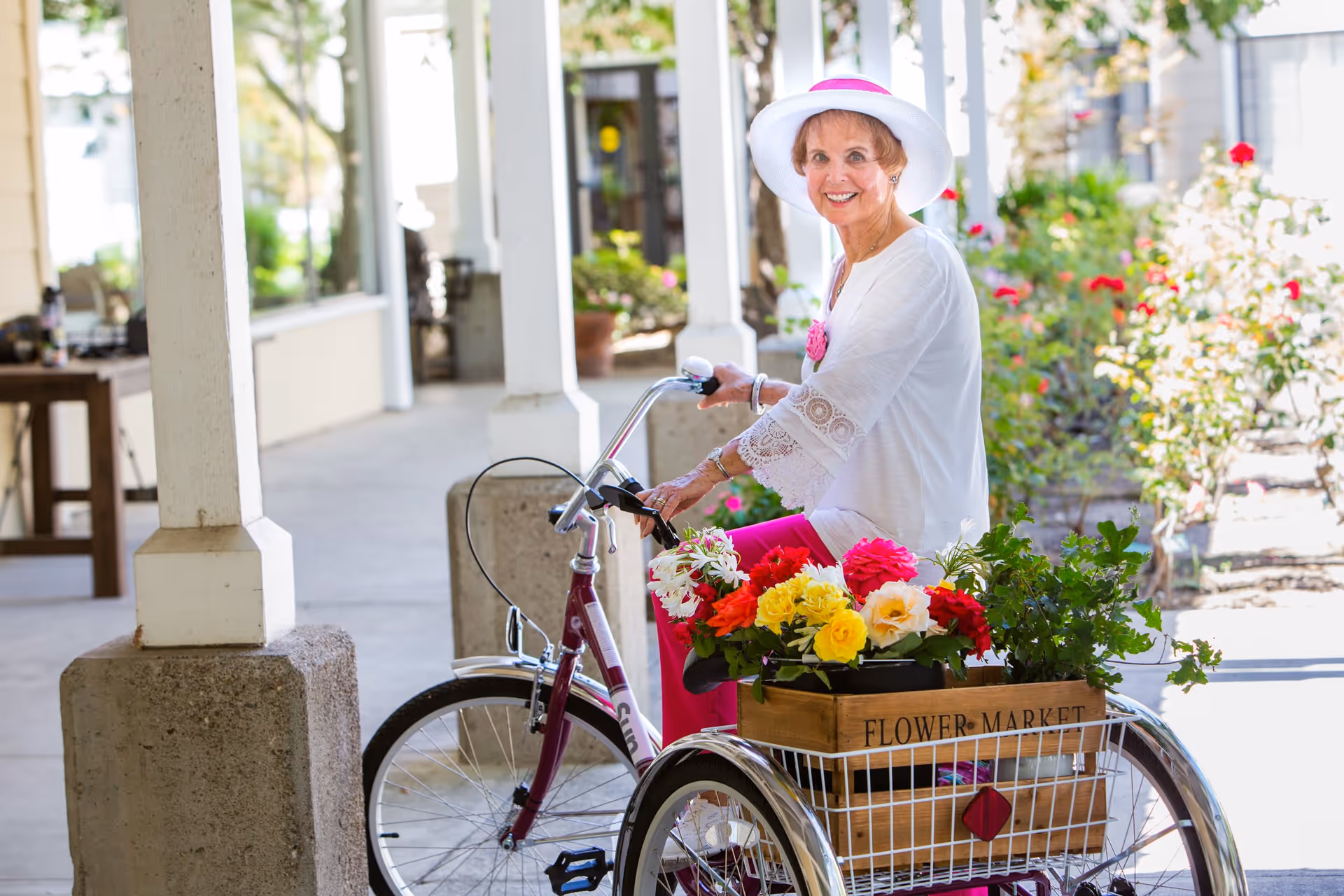 An elderly woman wearing a white hat and white blouse with lace sleeves sits on a purple tricycle with a wooden crate labeled 'FLOWER MARKET' filled with colorful flowers and greenery. She is smiling and outdoors near a building with white pillars and a garden with blooming flowers.