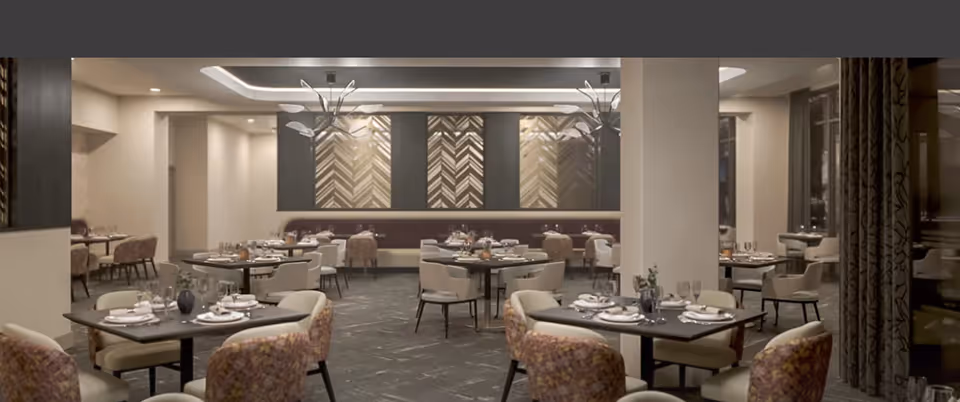 A modern dining room with multiple tables set for meals, featuring cushioned chairs in beige and patterned upholstery, decorative wall panels, and contemporary light fixtures hanging from the ceiling.