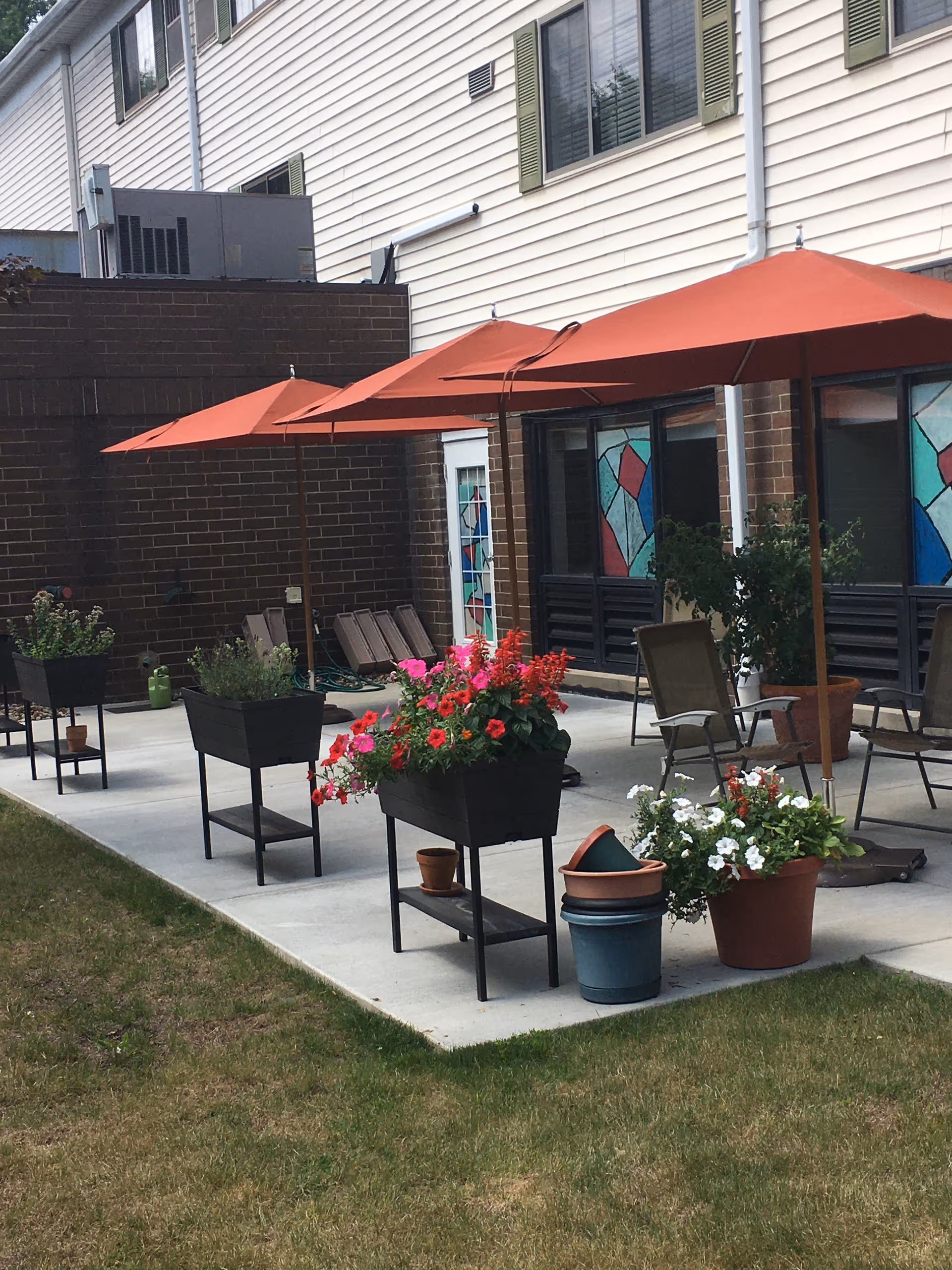 Outdoor patio area with several red umbrellas providing shade over chairs and tables. There are multiple planters with colorful flowers and greenery on a concrete surface next to a building with brick and siding exterior walls. Stained glass windows are visible on the building.