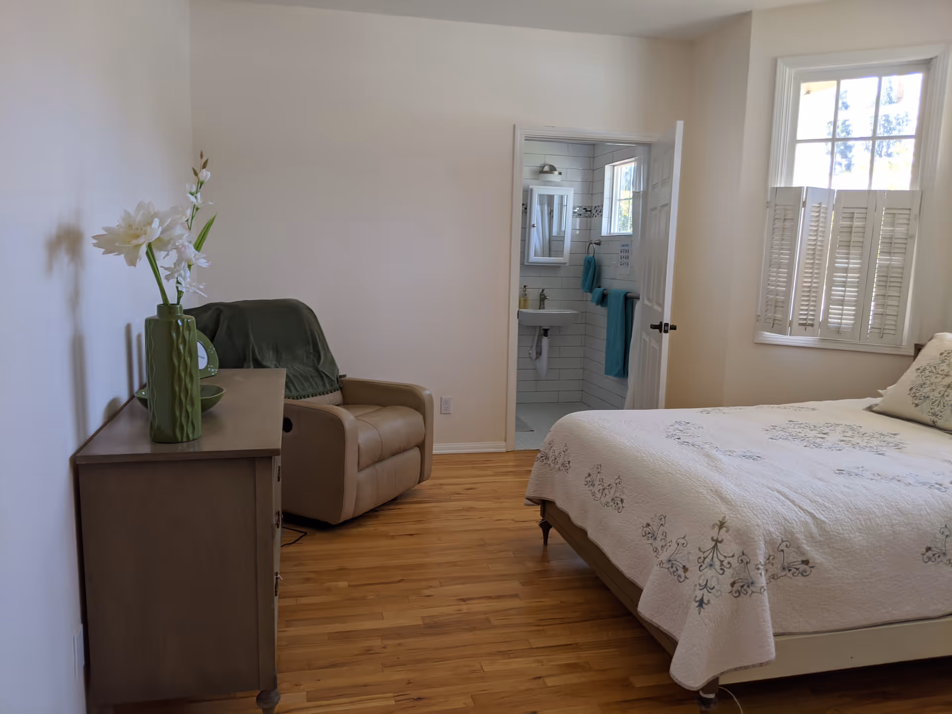 A bedroom with a wooden floor, a bed with a white and blue patterned quilt, a beige recliner chair with a green throw blanket, and a wooden dresser with a green vase holding white flowers. There is a window with white shutters and an open door leading to a bathroom with white tiled walls and teal towels.