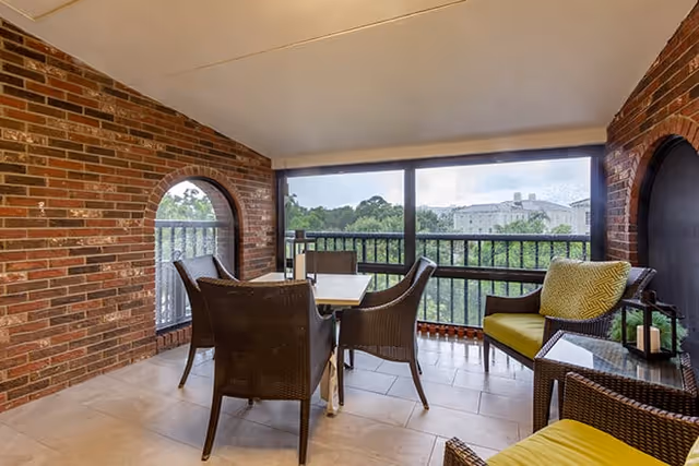 Covered outdoor patio area with brick walls and arched windows, featuring a table with four wicker chairs and a separate wicker seating area with yellow cushions and a glass-top side table. The patio overlooks greenery and a distant building.